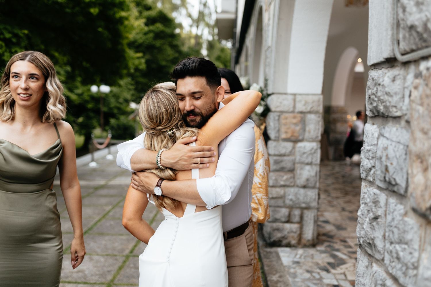 Wedding party members in olive green and white attire posing together against stone wall backdrop.