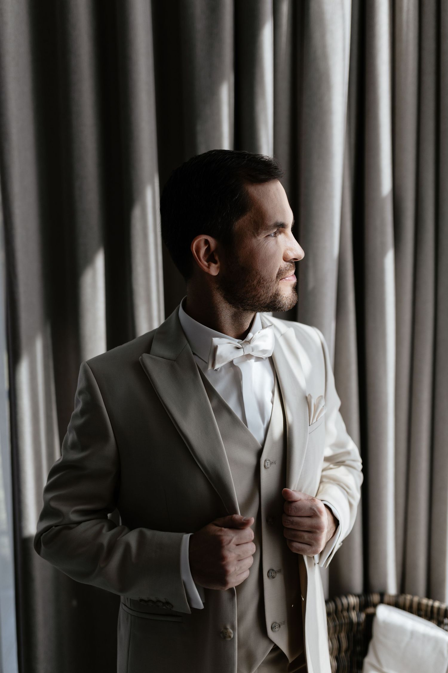 A groom in a light gray suit and white bowtie adjusts his jacket while standing by curtained windows.