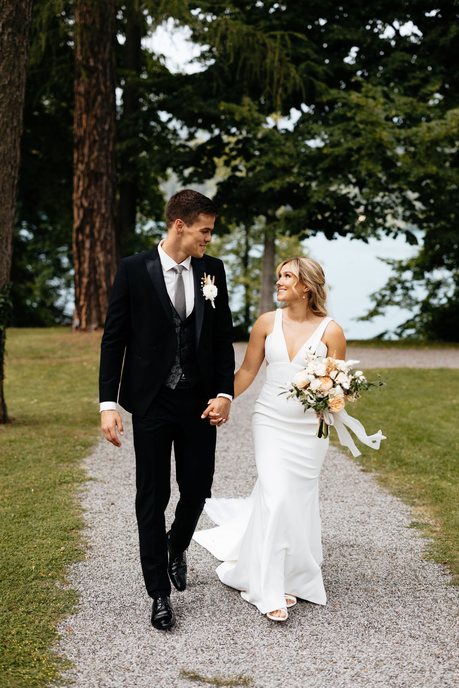 A couple in wedding attire walks hand in hand down a gravel path surrounded by tall evergreen trees.