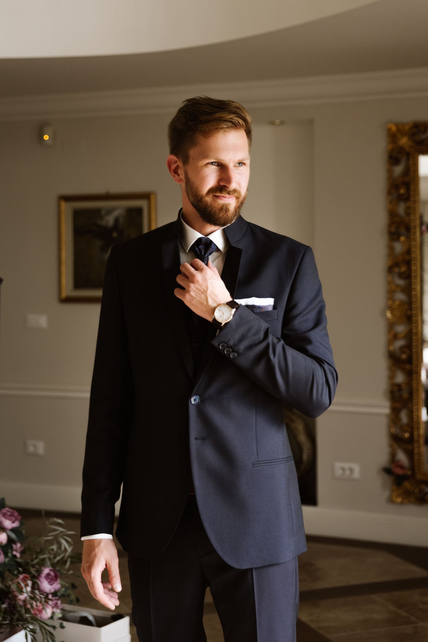 A groom adjusts his tie while standing in a formal navy suit against an elegant interior backdrop.