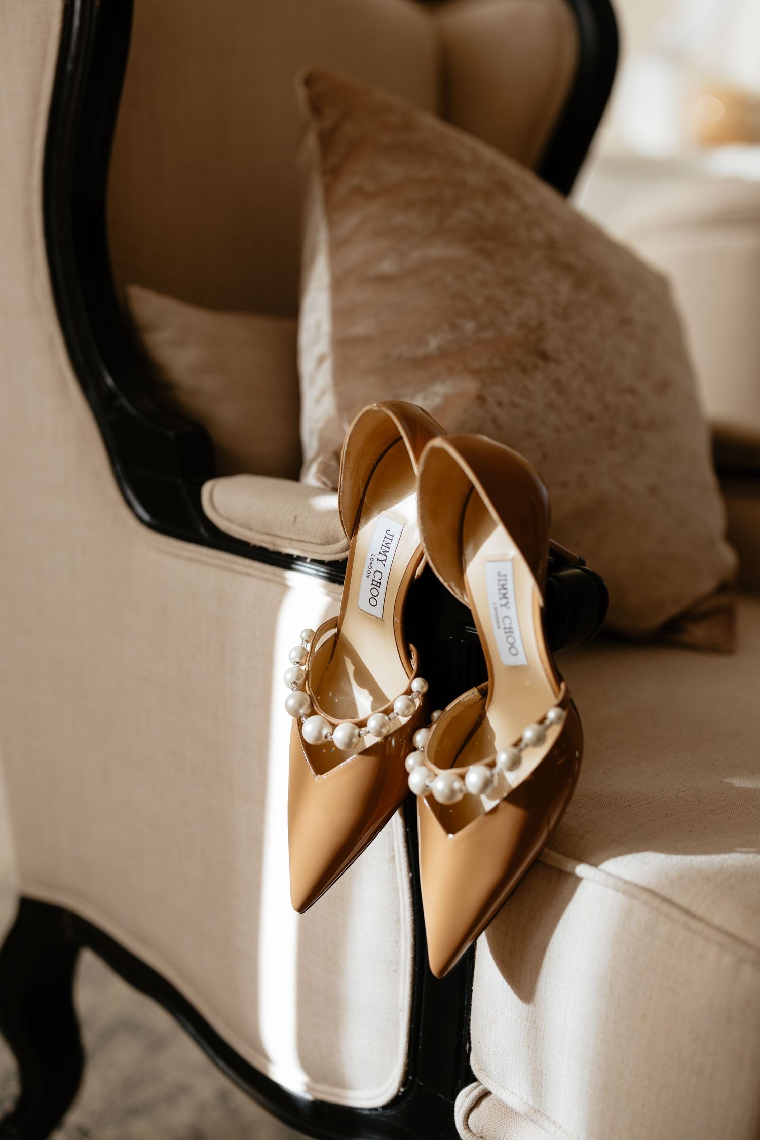 Elegant beige leather bridal shoes with pearl embellishments sit in soft window light on a white surface.
