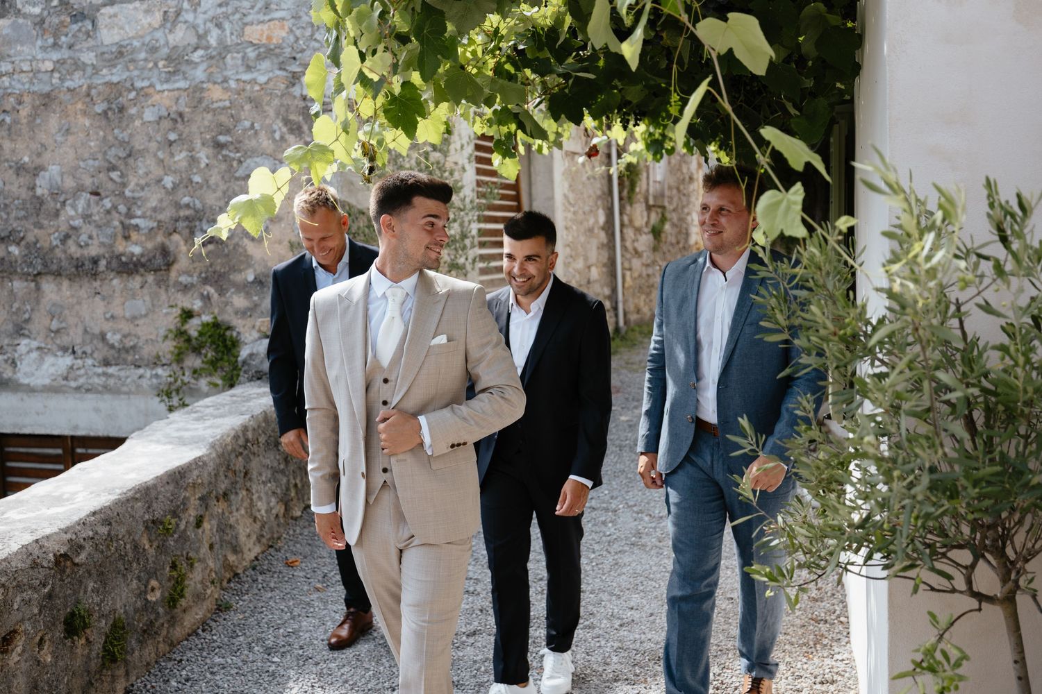 Wedding party members walk together on a stone path outside a Mediterranean-style building.