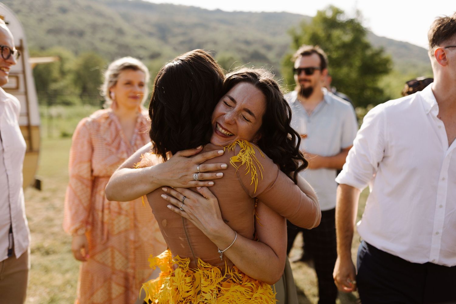 Guests share emotional embraces during an outdoor celebration with rolling hills in the background.