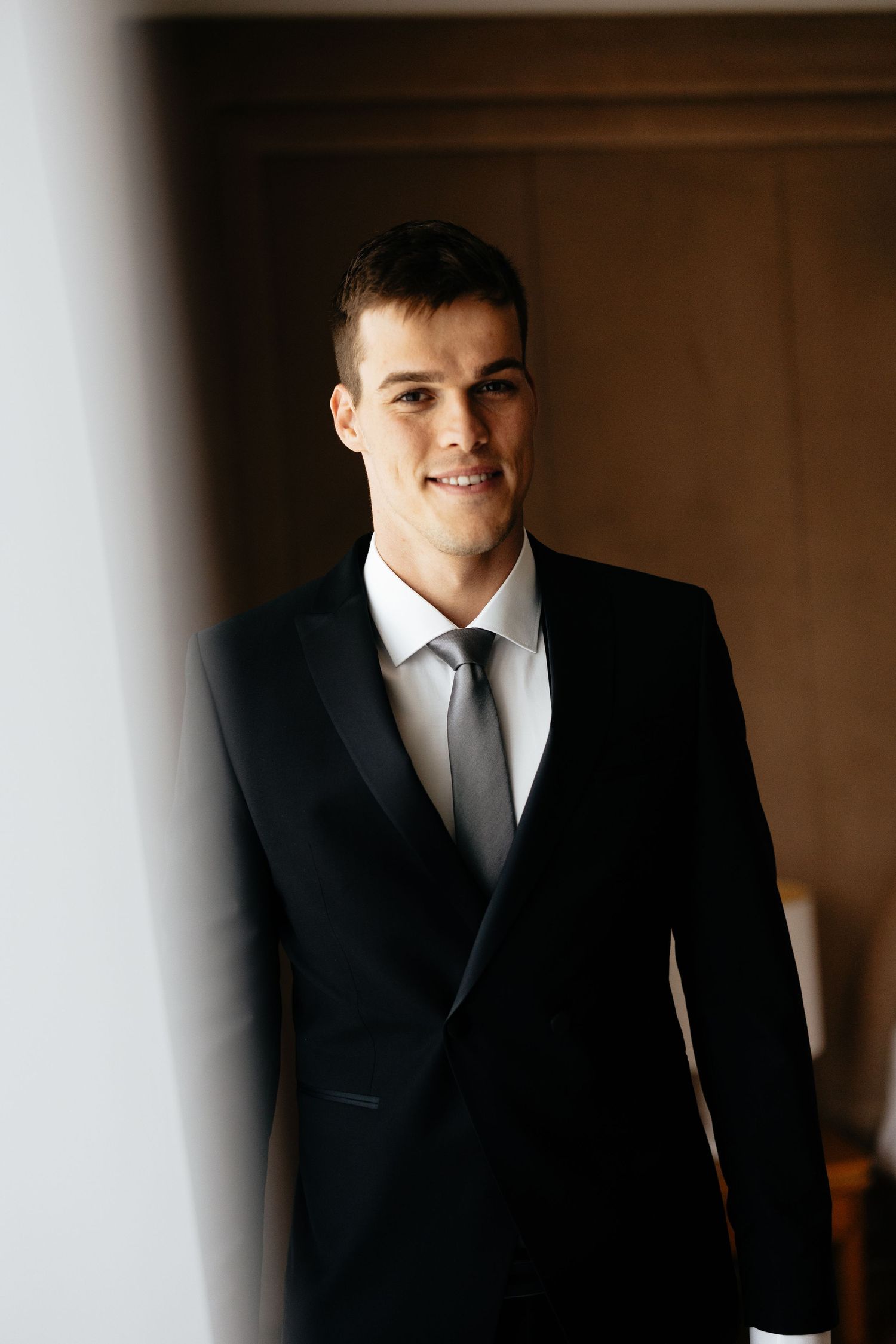 A groom in a classic black suit and tie stands in a dimly lit room before his wedding ceremony.