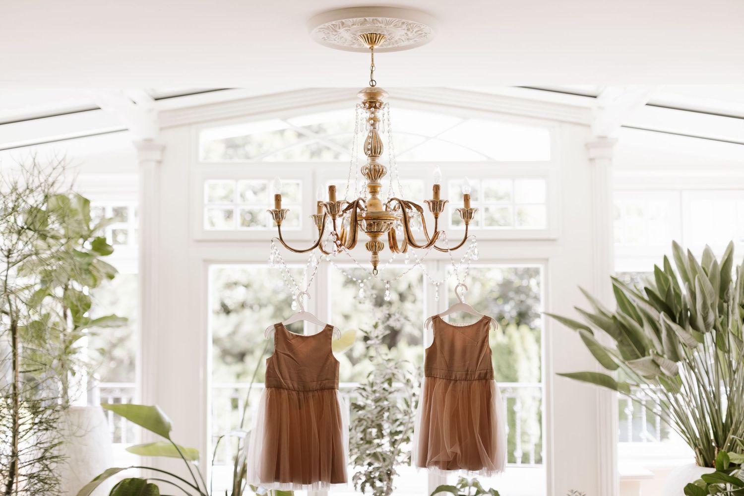 Elegant brown bridesmaid dresses hanging in a bright white room with chandelier and natural light.