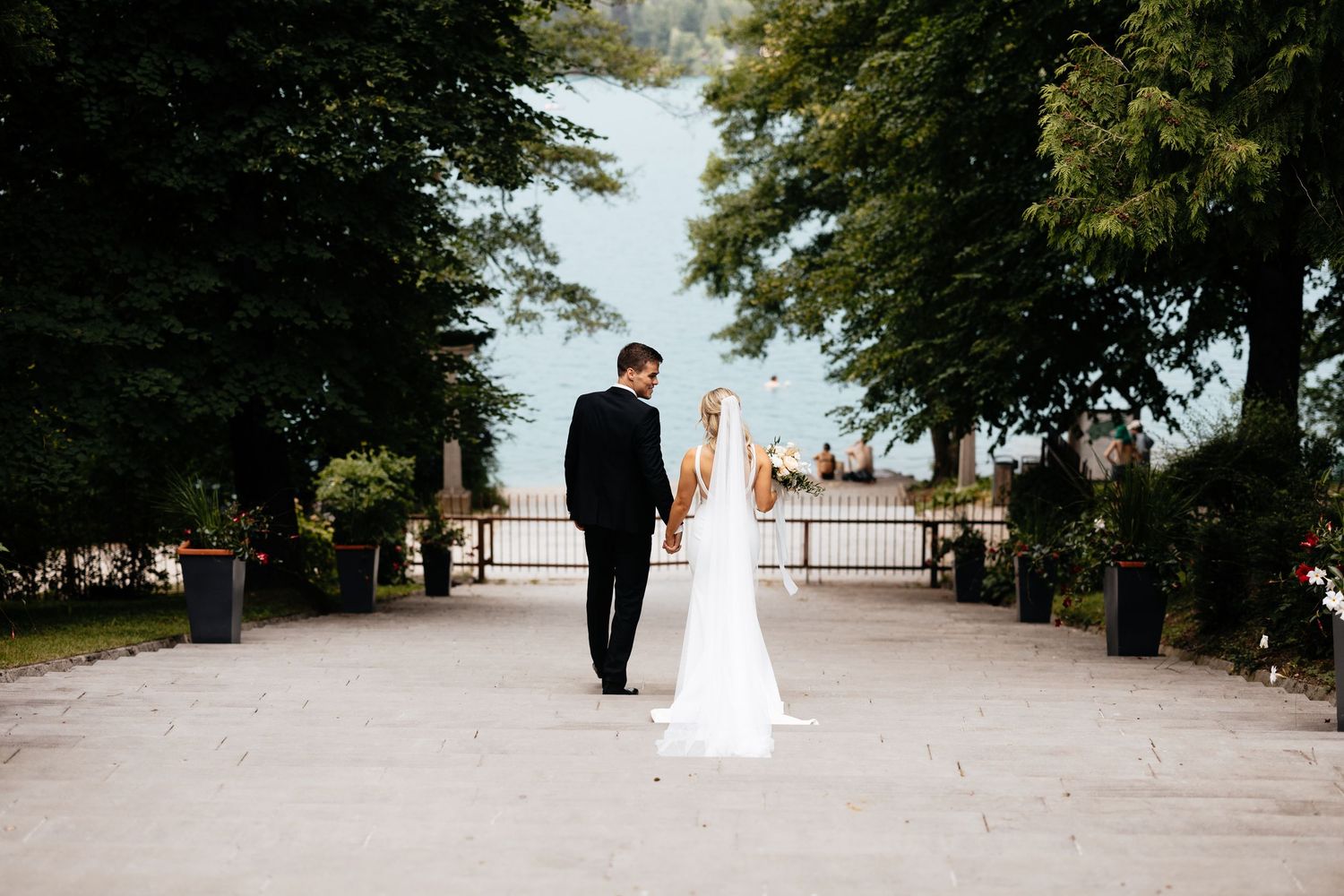 A couple in wedding attire walking down a tree-lined path towards a scenic overlook on their special day.