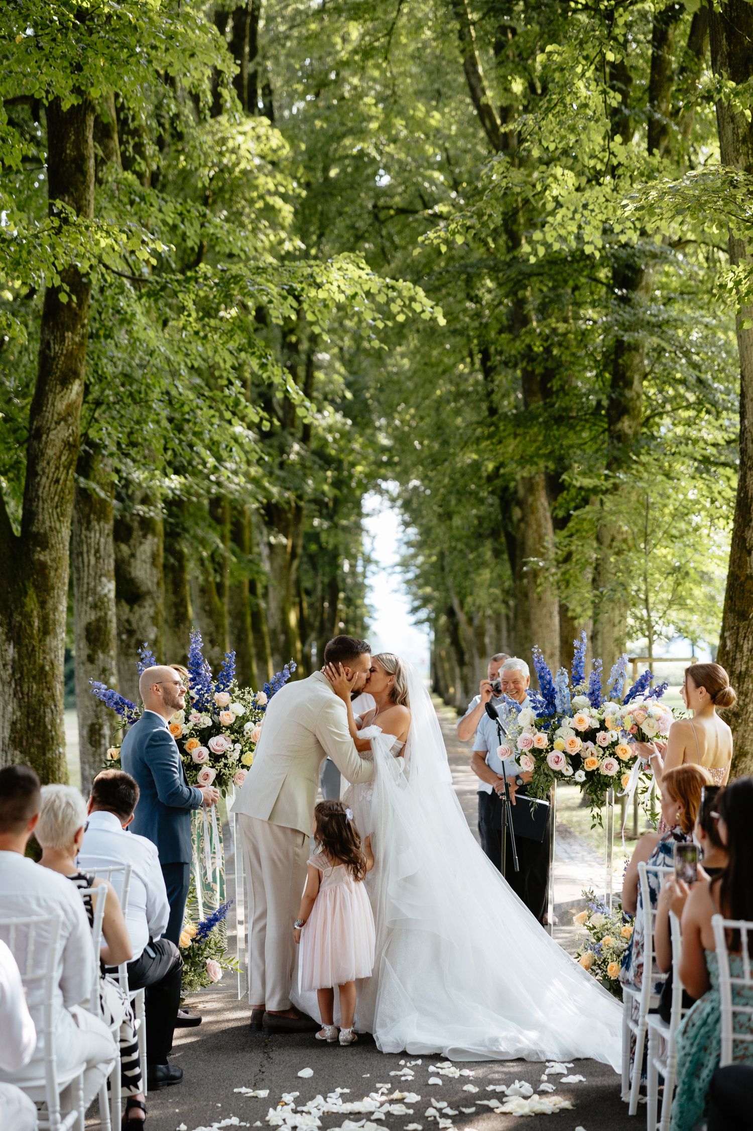 A romantic outdoor wedding ceremony takes place under a tree-lined path with white chairs and floral arrangements.