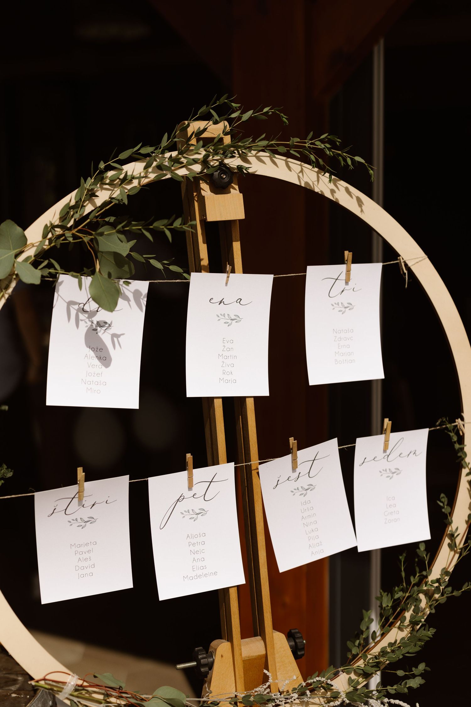 Wedding seating chart cards displayed on string with greenery decorations and wooden arch.