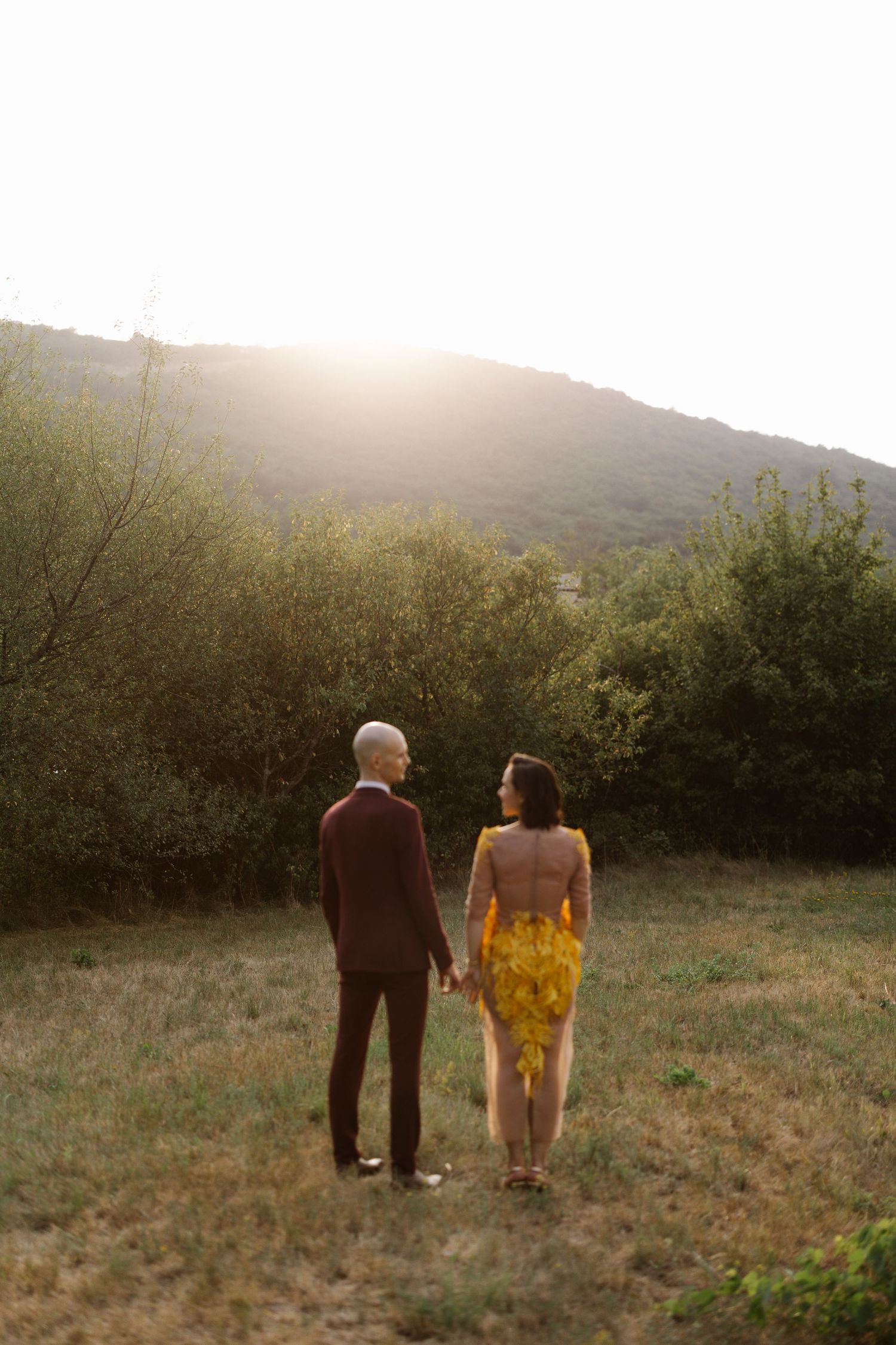 Romantic couple walking hand in hand through a sunlit meadow with rolling hills in the distance.