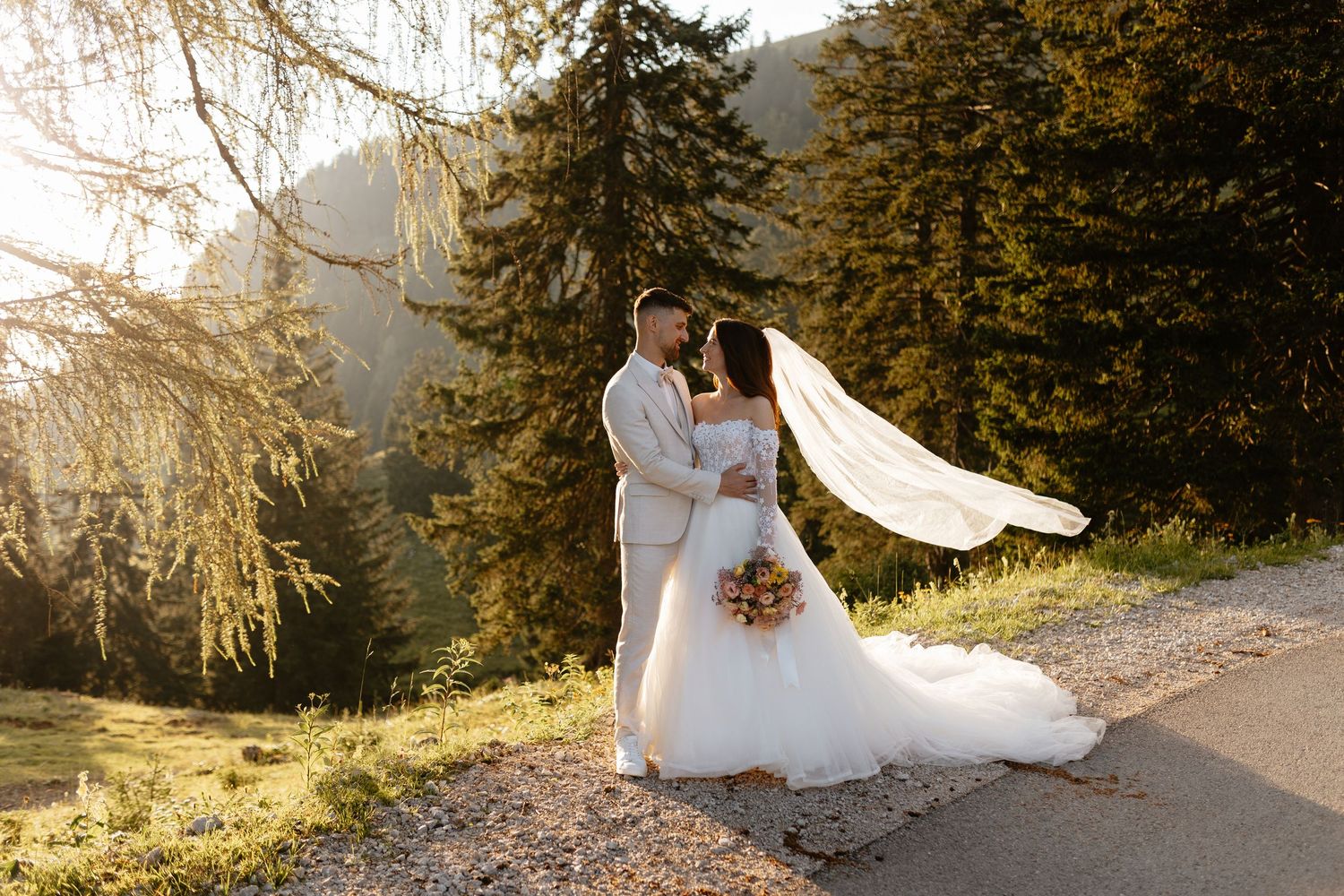 Wedding couple poses on a scenic mountain trail at sunset with the bride's veil flowing in the breeze.