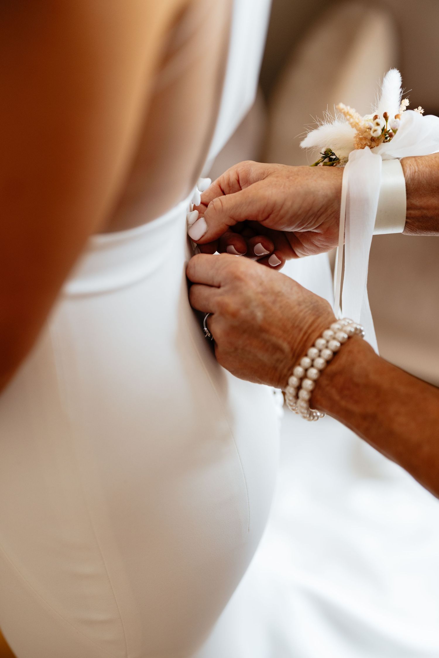 Hands wearing pearl bracelets adjust the back of a white wedding dress.