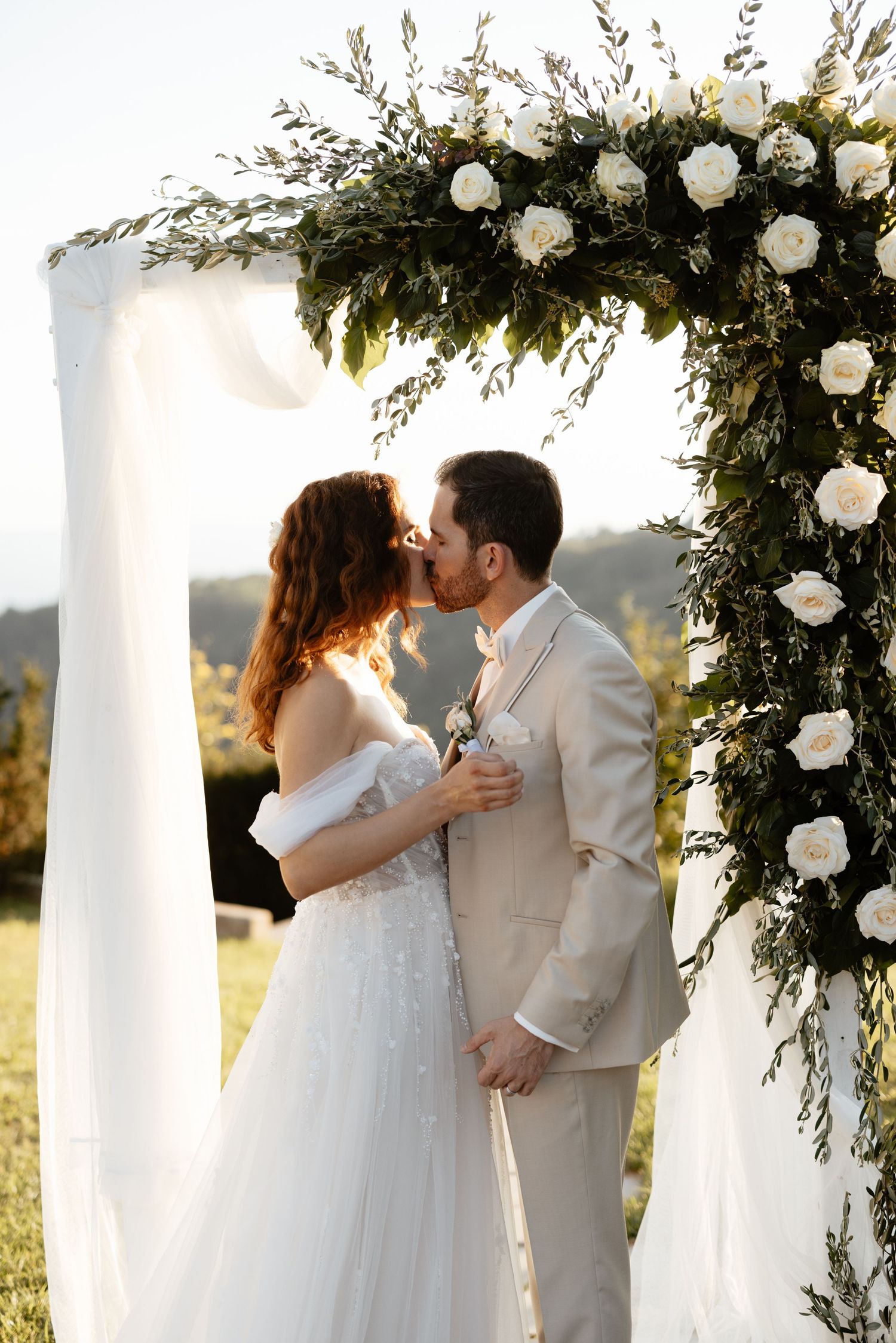 Wedding couple embraces under white pillars decorated with roses and greenery during outdoor ceremony.