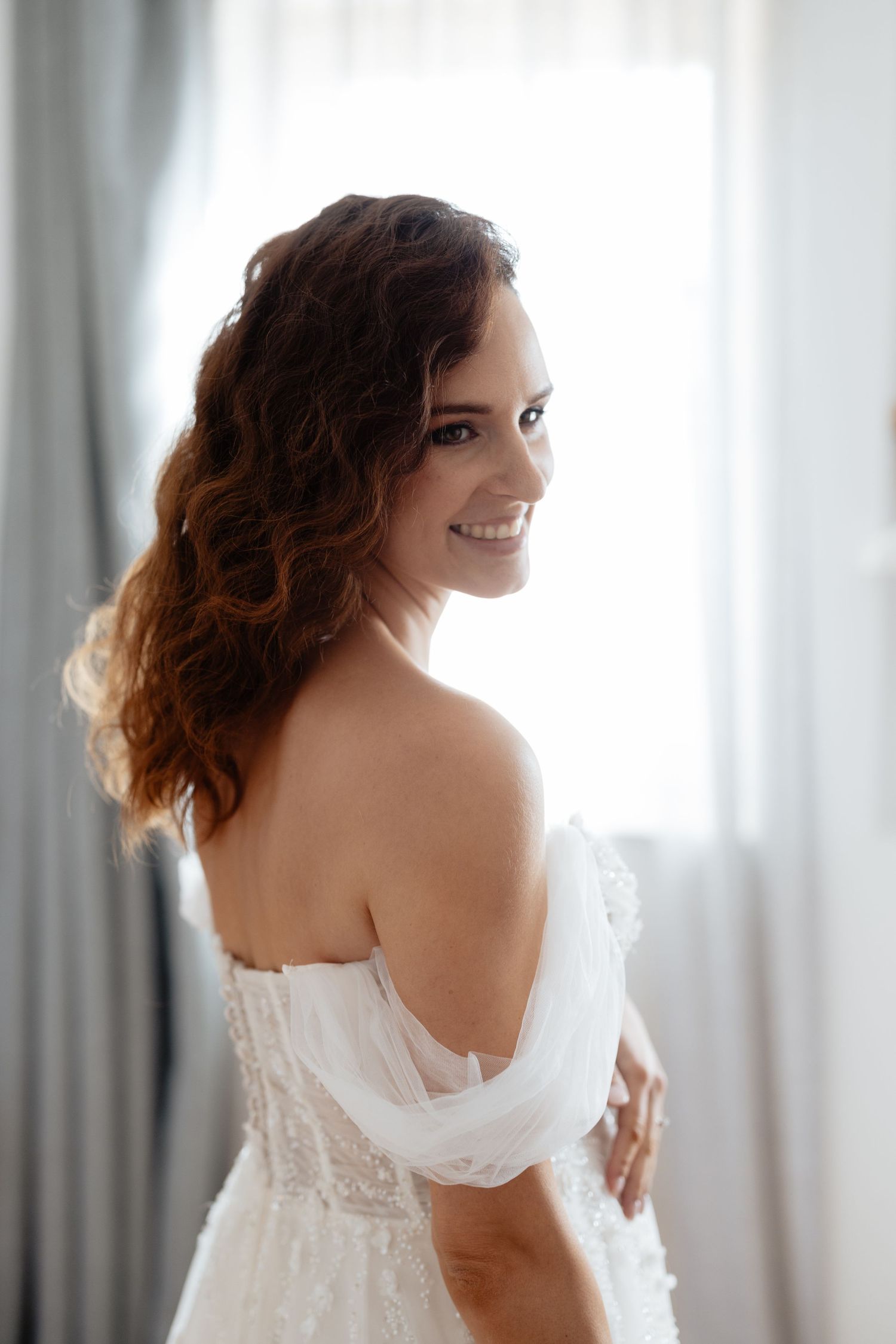 Bride in white wedding dress poses gracefully by window light in intimate boudoir style photoshoot.