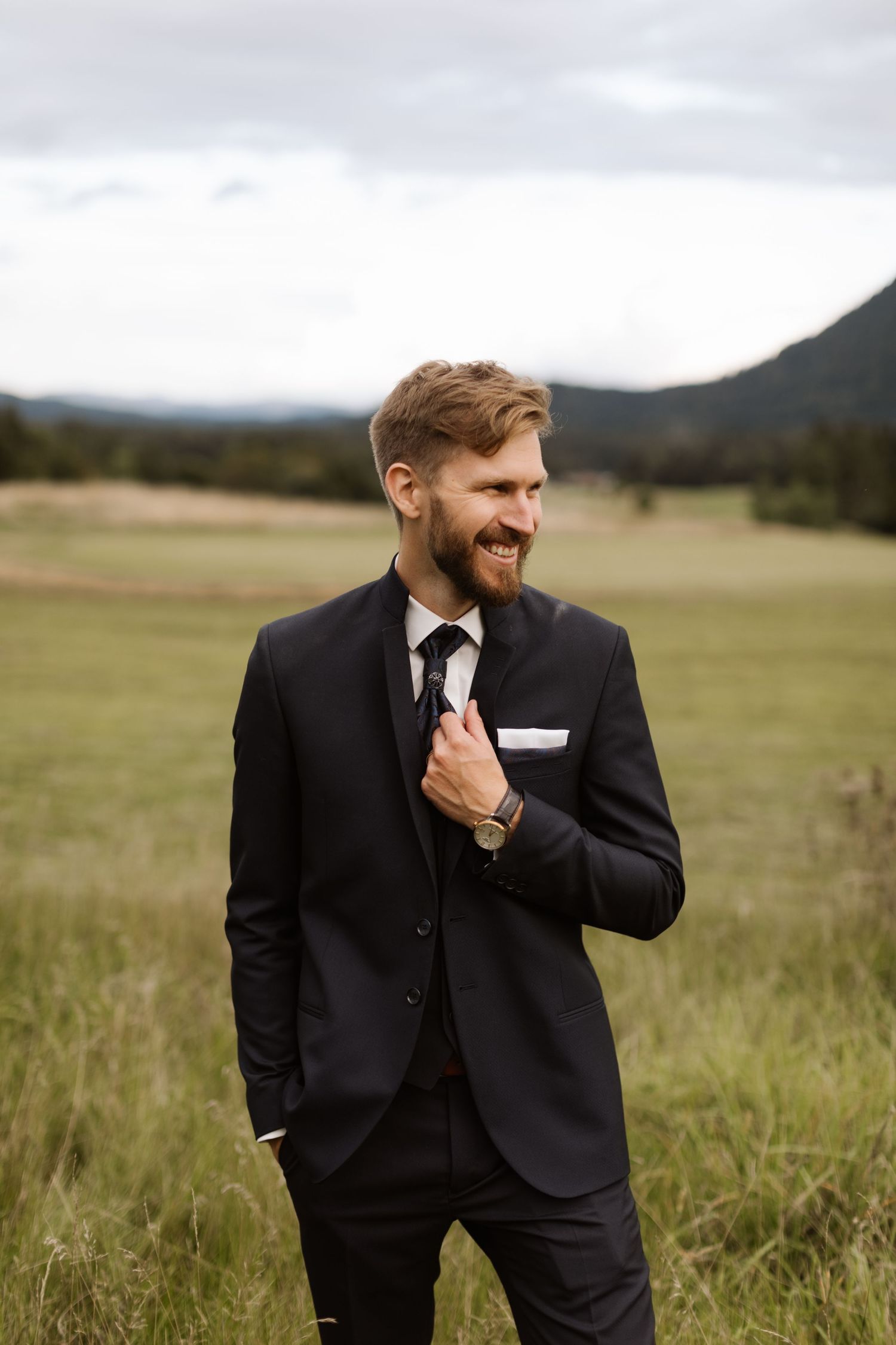 Groom in classic black tuxedo laughing joyfully in an outdoor field setting.