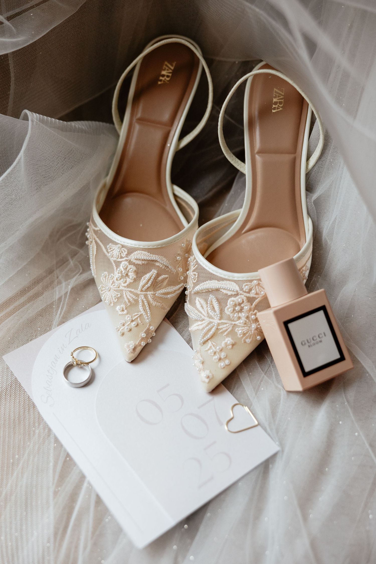 Elegant white bridal shoes with floral details displayed with wedding rings and a perfume bottle.