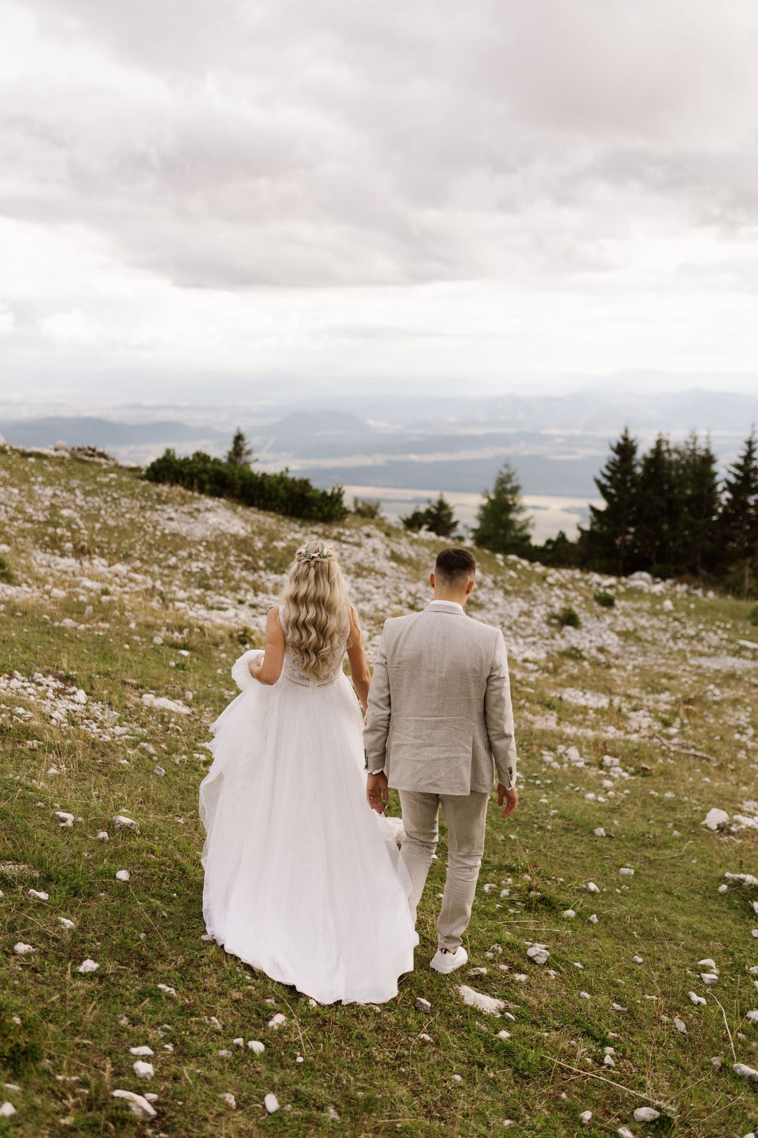 A couple in wedding attire walks together on a mountainside overlooking a misty valley.