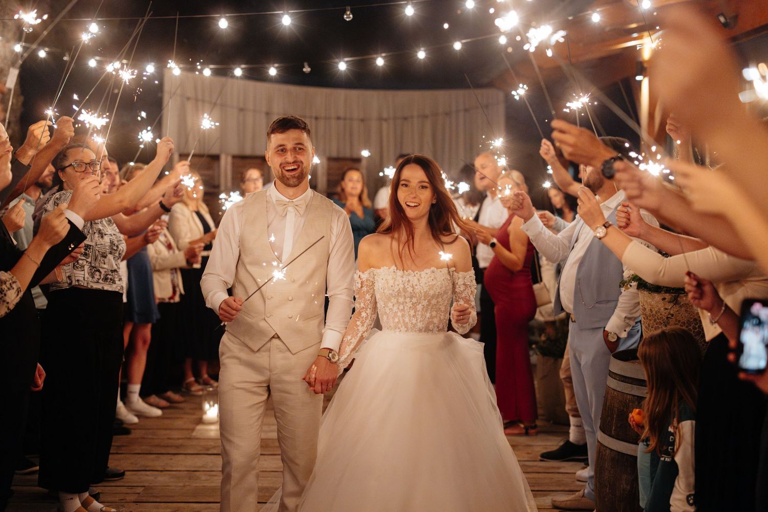 Wedding reception guests dance and celebrate under twinkling string lights at an indoor venue.