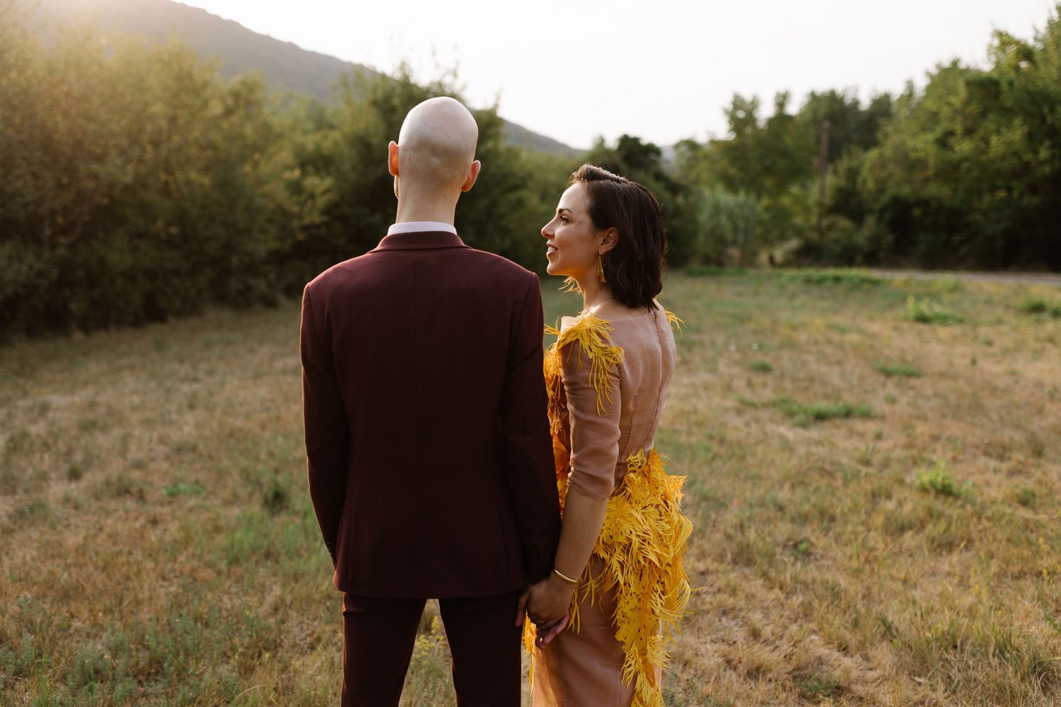 A couple stands together in a golden sunset field with mountains in the background.