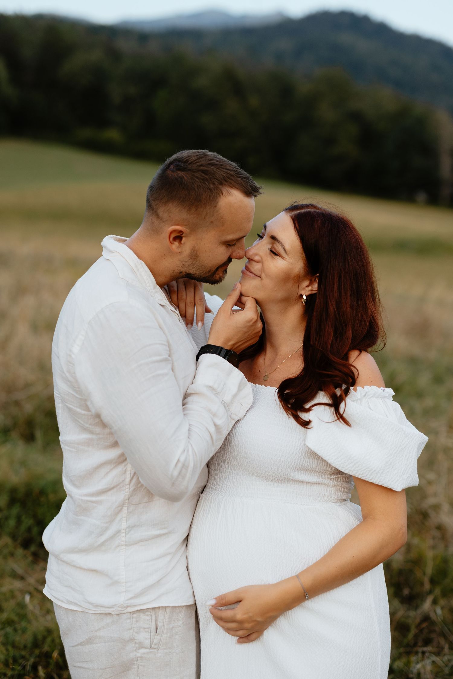 A romantic pregnancy photoshoot of a couple in white outfits standing in a mountain field.