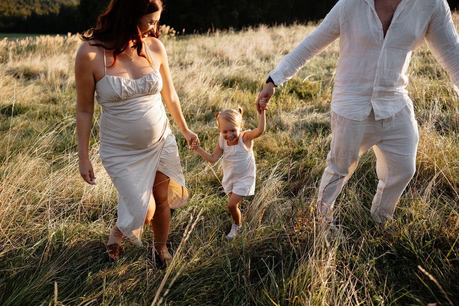 Couple in all-white outfits walking playfully through a sunlit field of tall grass.