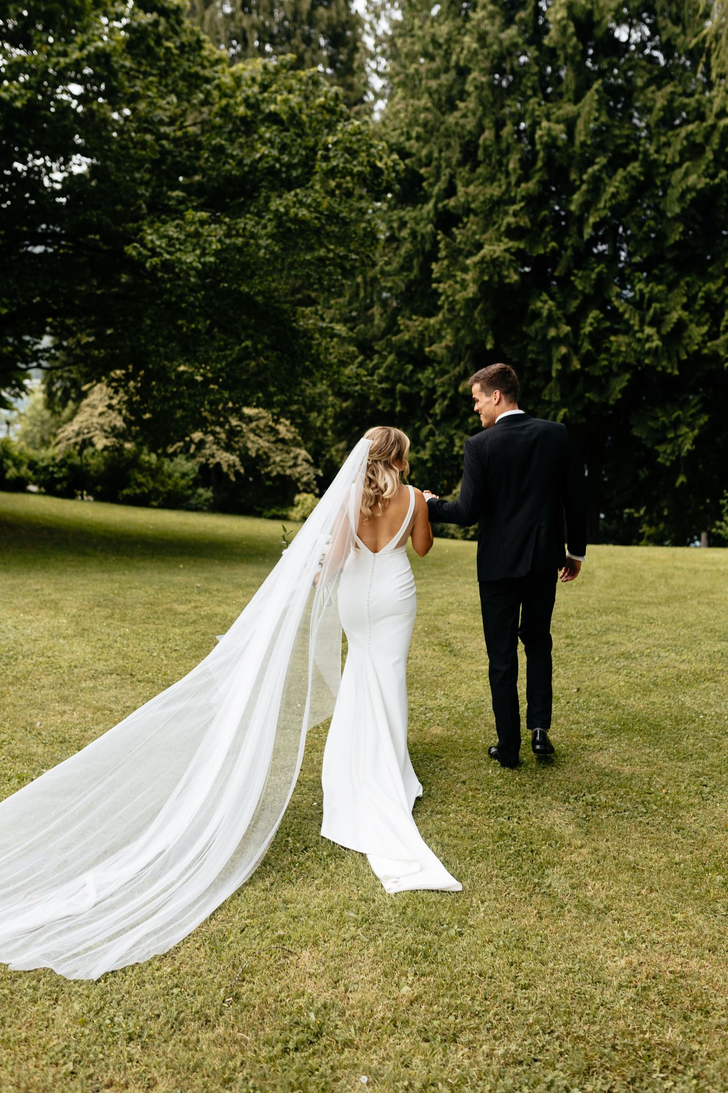 Long flowing white wedding dress train trailing behind couple on green lawn.