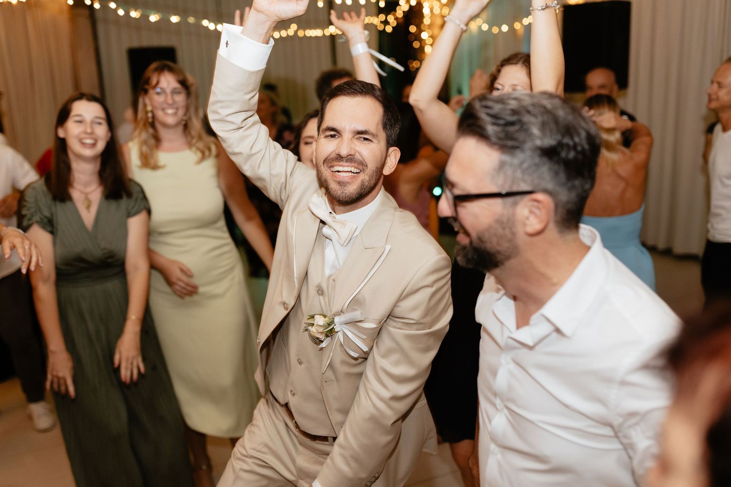 Wedding guests dancing and celebrating on the dance floor with string lights glowing overhead at an indoor reception.