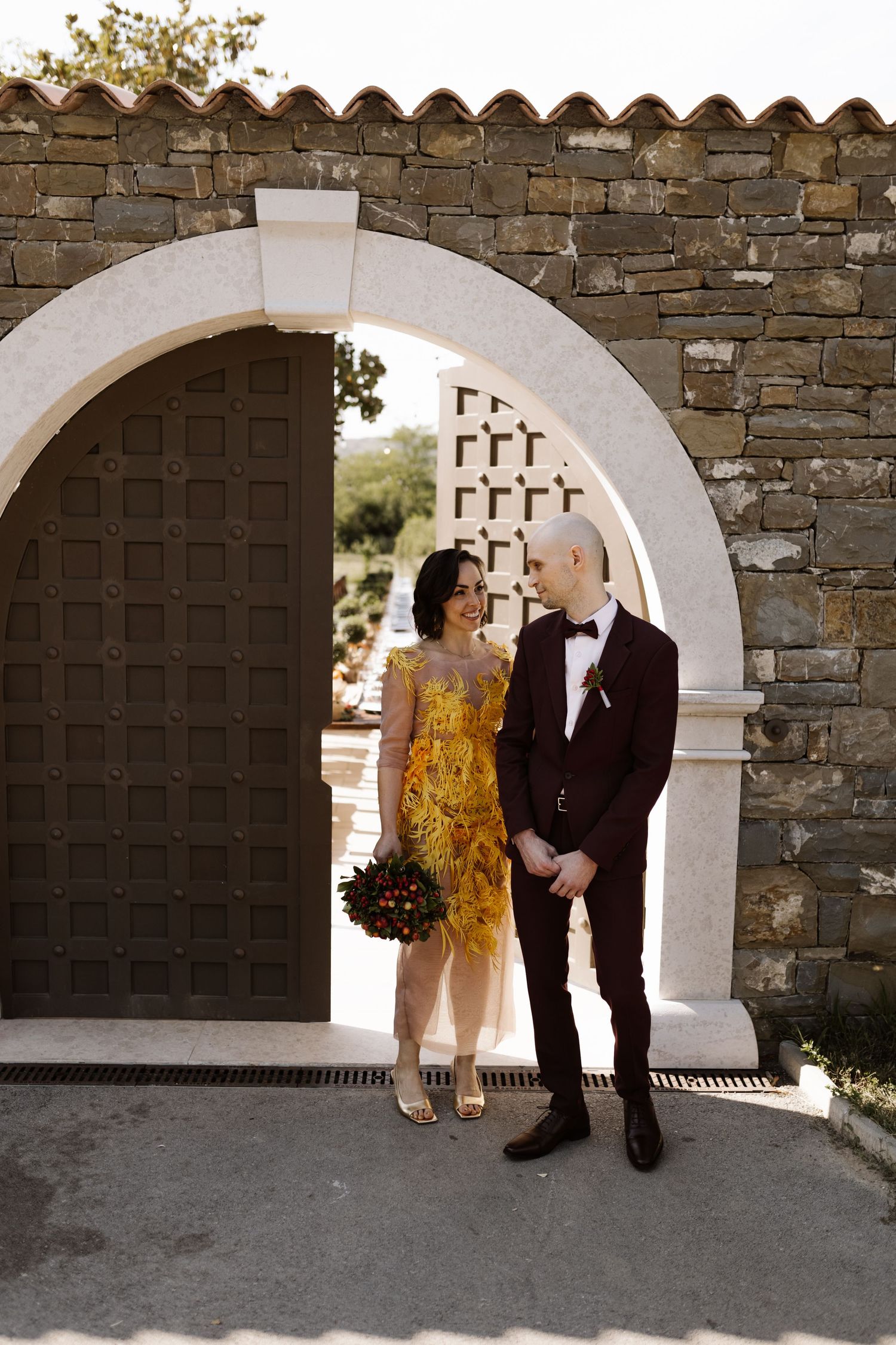 A couple in wedding attire stands beneath a stone archway, with the bride wearing a striking yellow sequined dress.