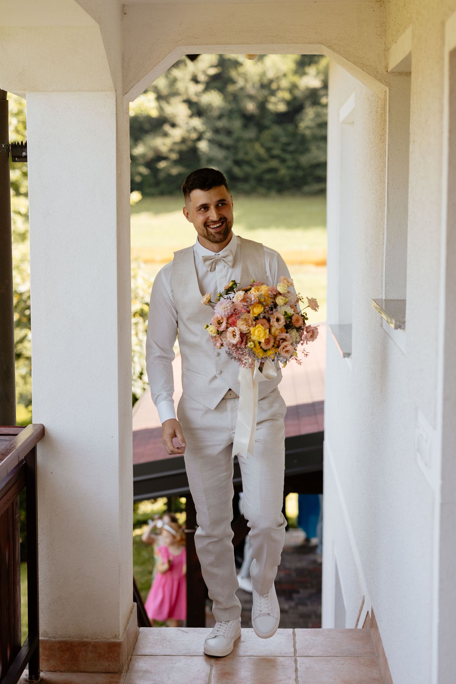 Person in white suit walking along a columned porch carrying a colorful bouquet.