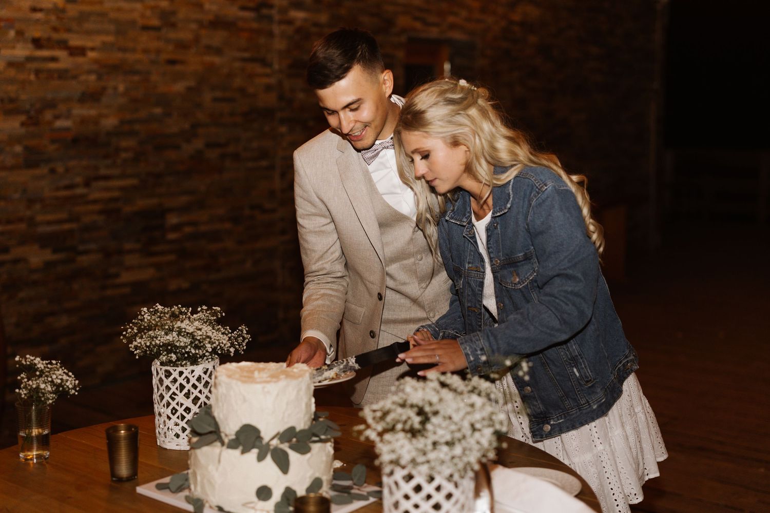A couple cuts their elegant two-tier wedding cake decorated with greenery at a rustic reception.