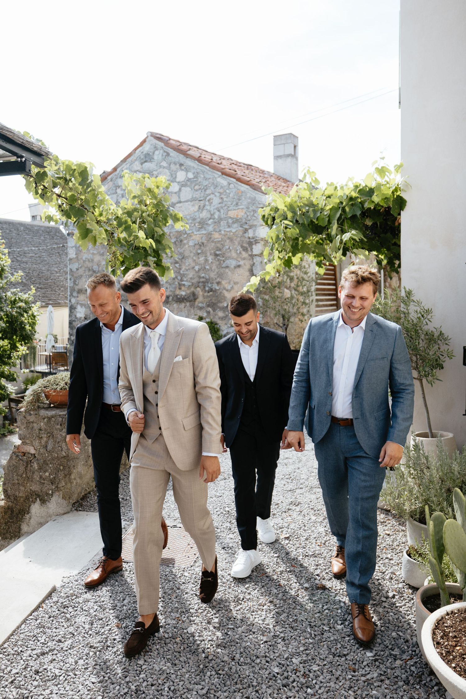 Groomsmen in mixed formal attire walking along a gravel pathway at an outdoor venue.