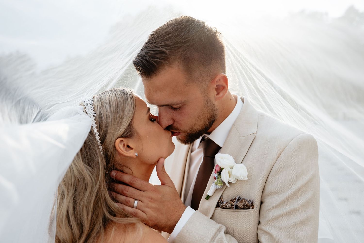 Romantic wedding couple sharing intimate kisses under flowing white veil outdoors.