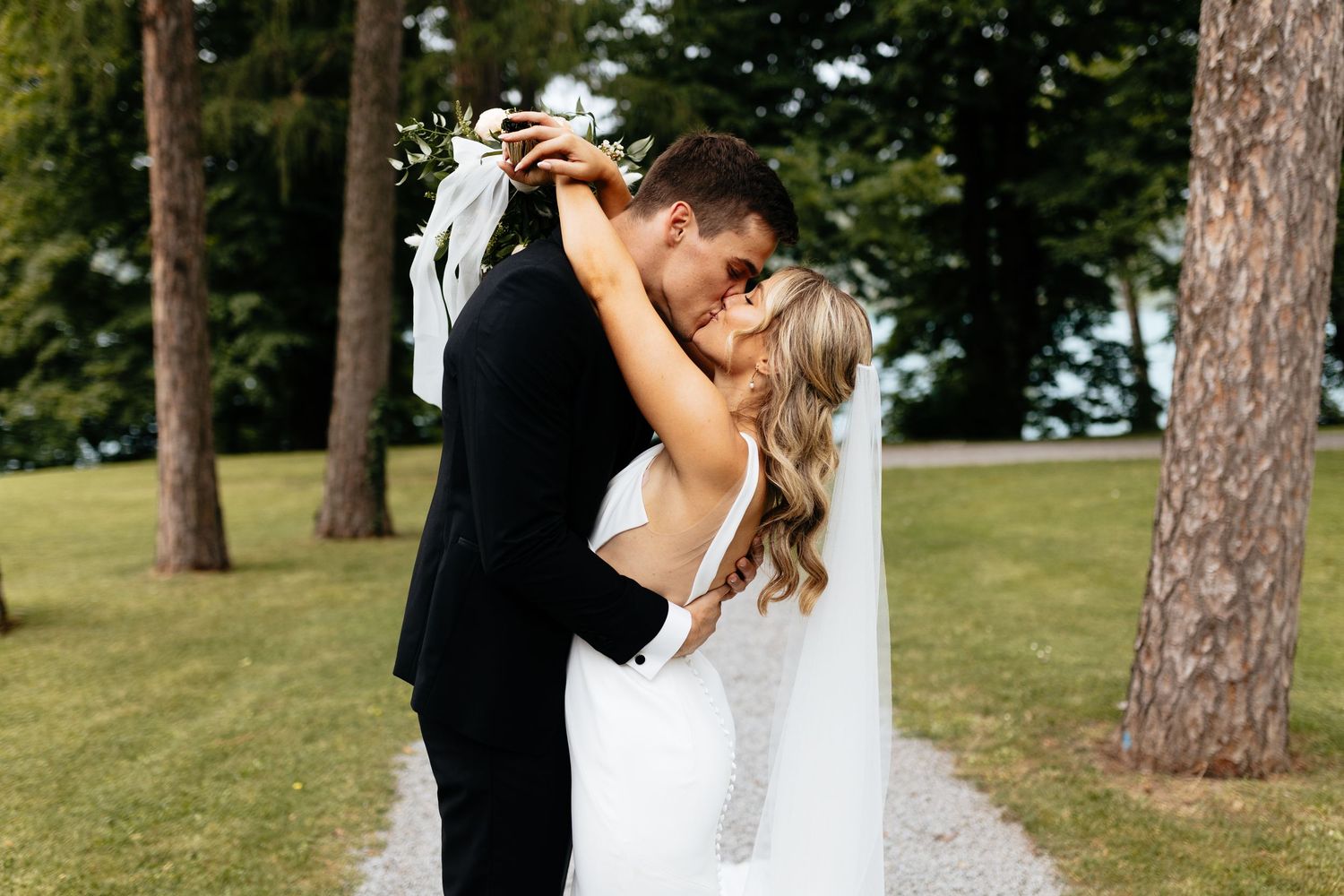 A romantic wedding couple embraces outdoors in a sunlit park with tall pine trees in the background.