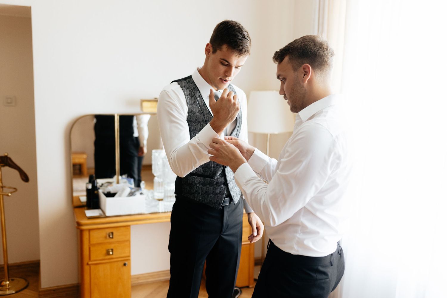 Two people in formal attire help each other get dressed in a sunlit room with wooden furniture.