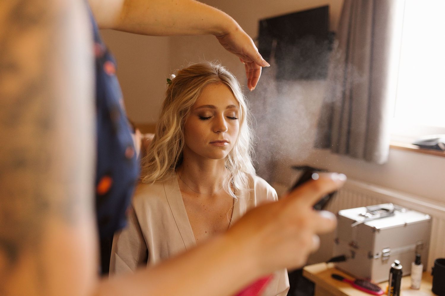 A bride getting her makeup professionally applied during wedding preparations in a softly lit room.