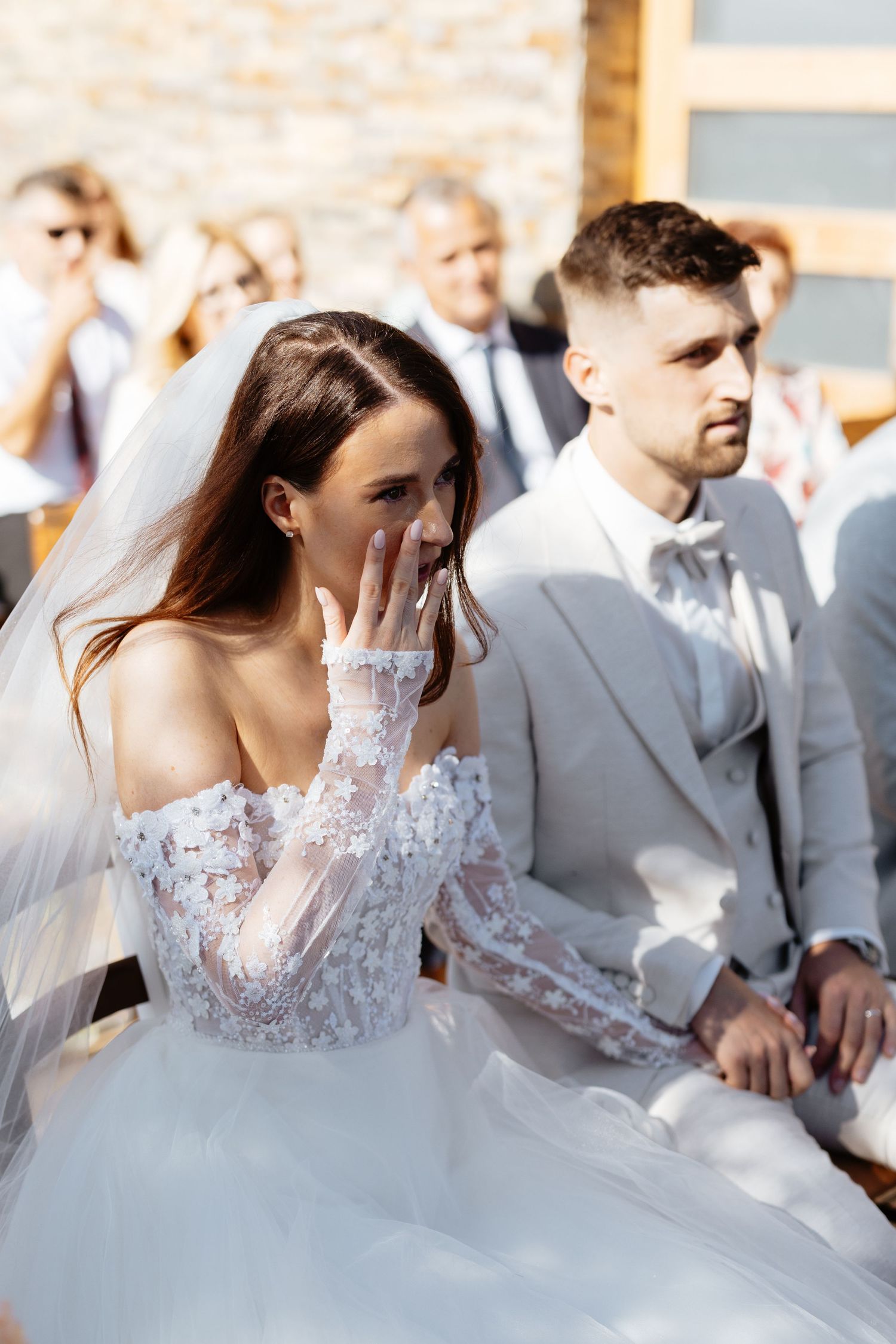 Wedding ceremony scene with bride in lace gown surrounded by guests in outdoor setting.