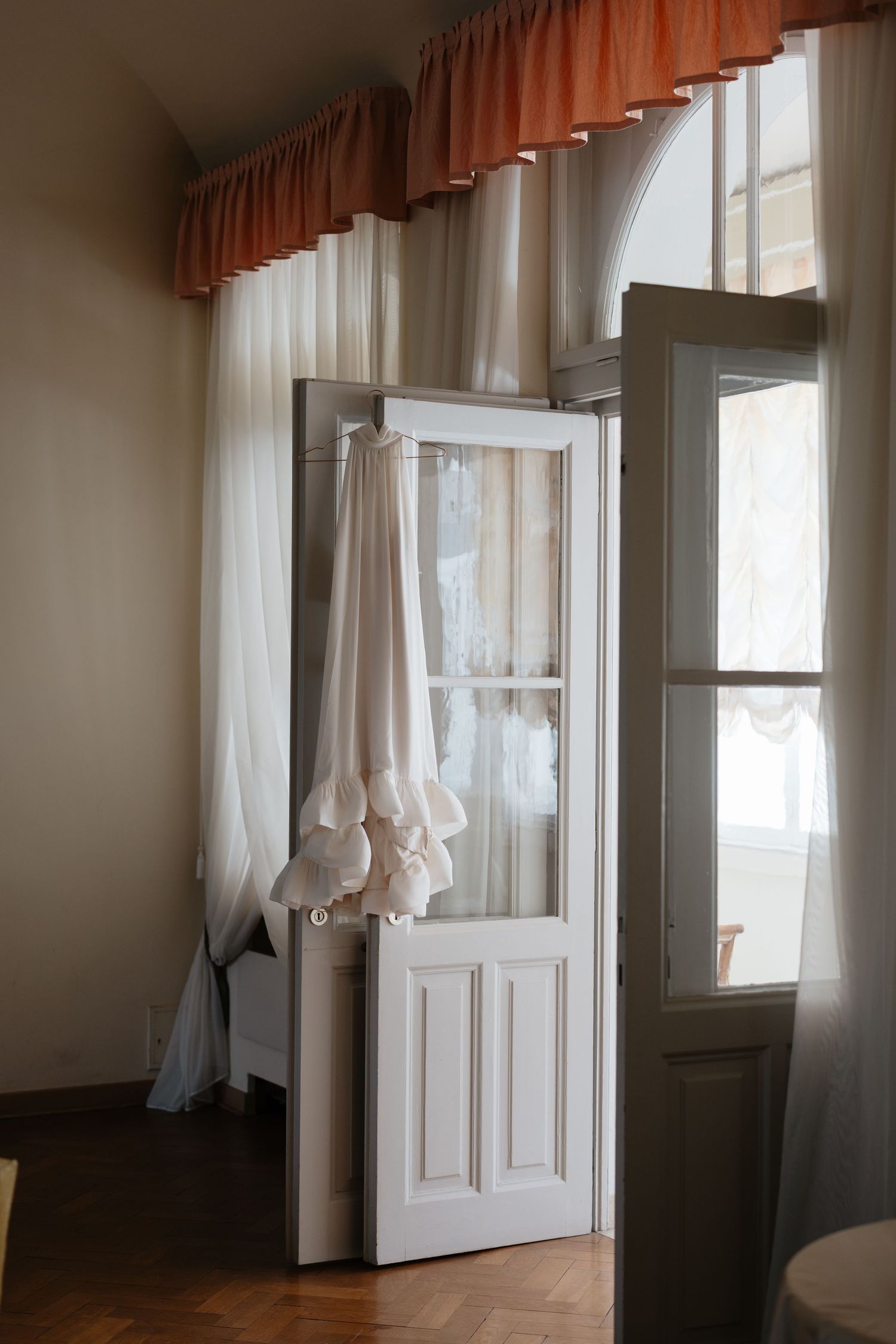 A white wedding dress hangs elegantly by a window with peach and white curtains.