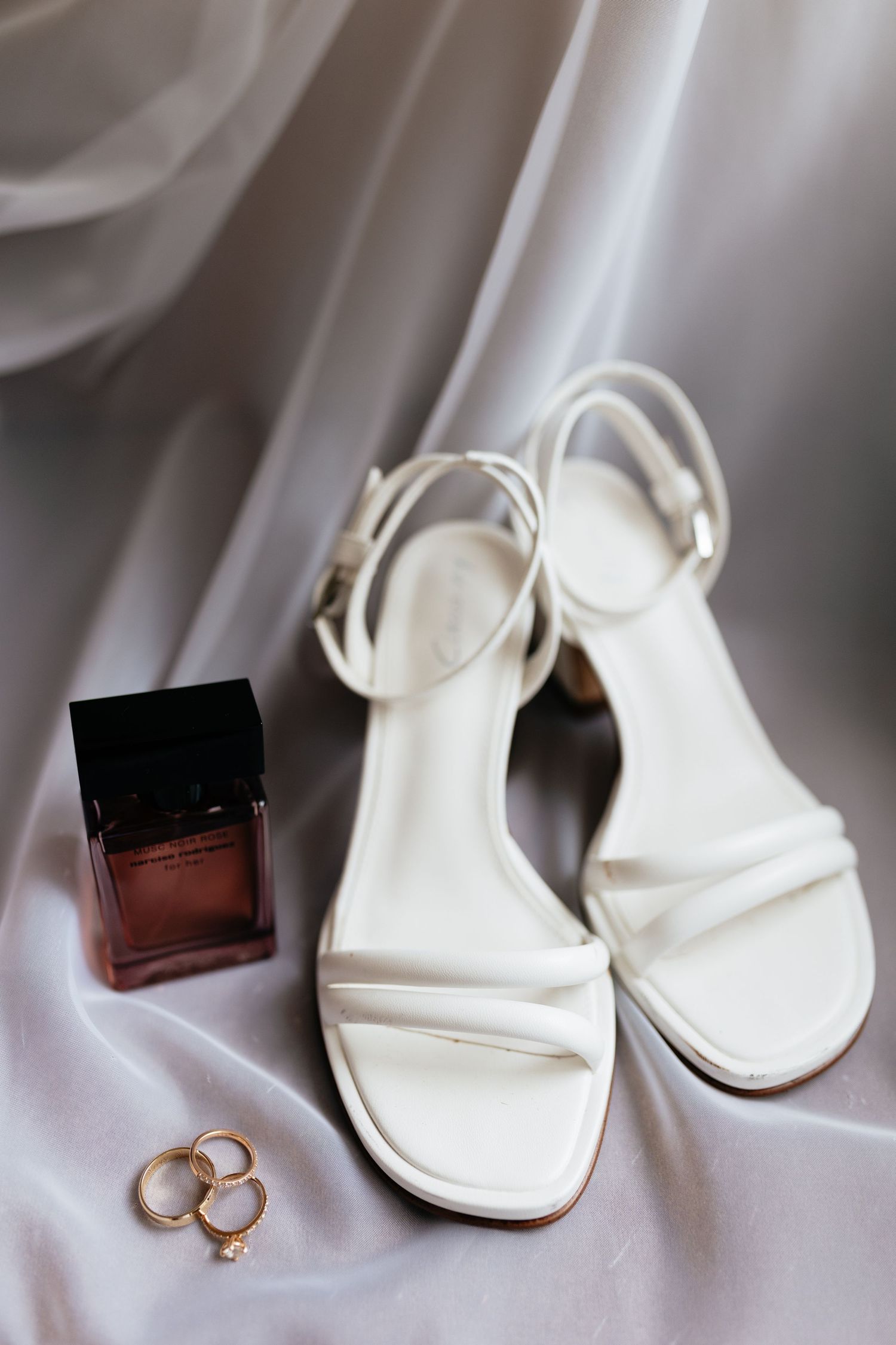 White bridal sandals and wedding rings arranged on silky fabric.