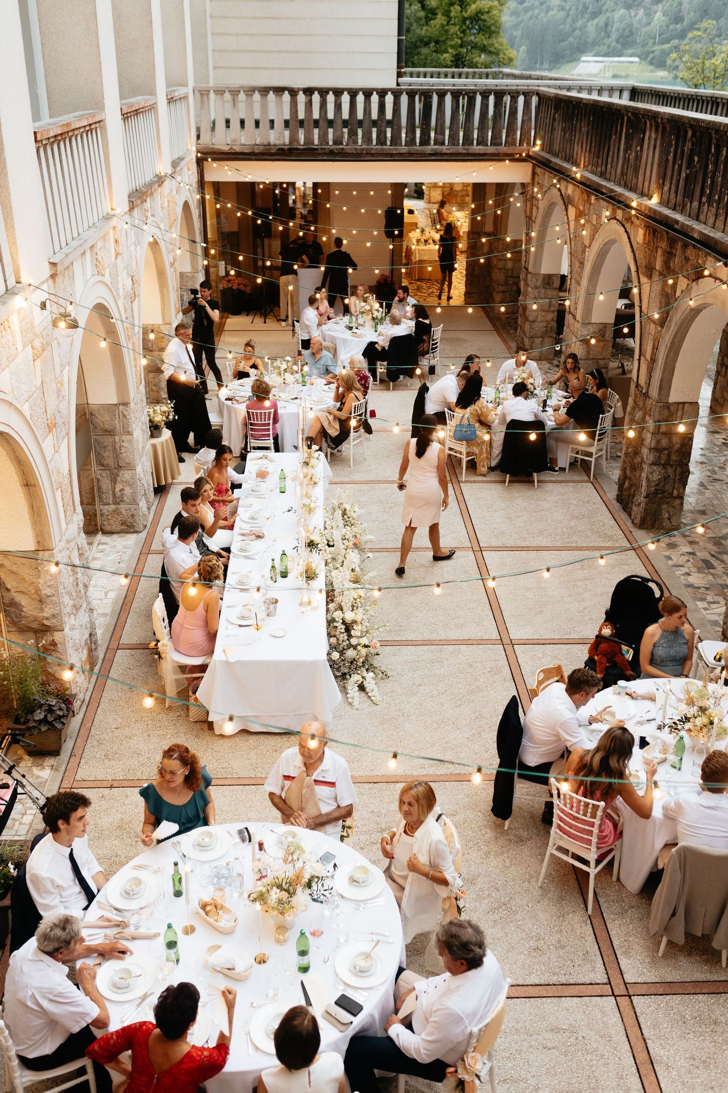 Wedding guests enjoy dinner at decorated tables under string lights in an outdoor courtyard.