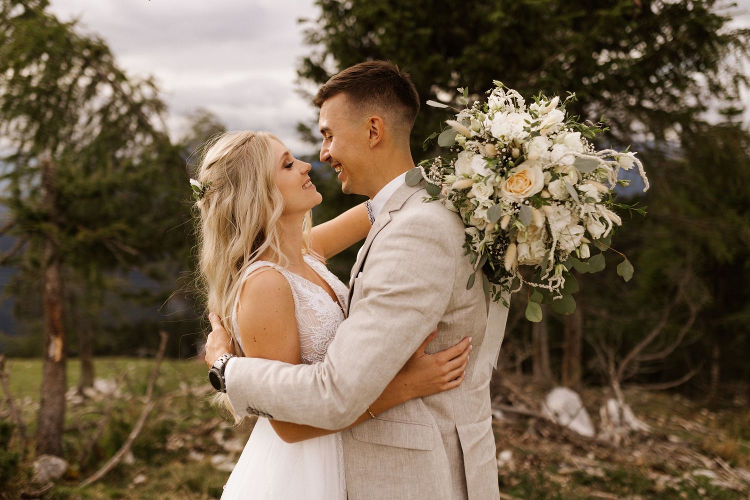A romantic wedding couple embraces outdoors surrounded by lush greenery and white floral bouquets.