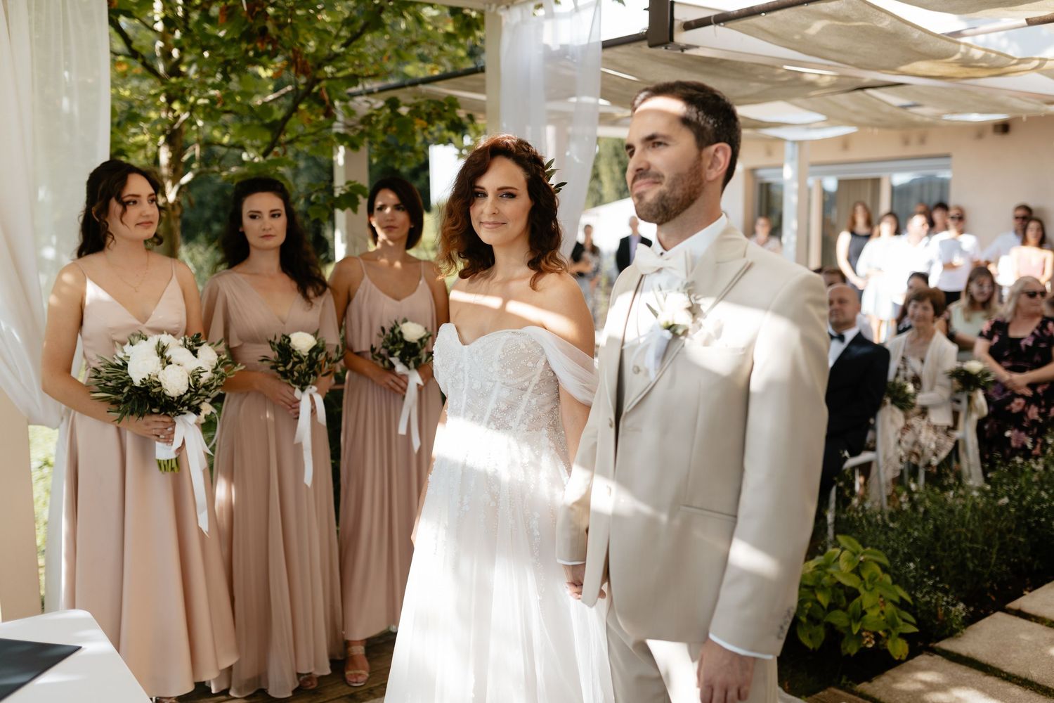 Bridesmaids in elegant champagne-colored dresses stand together at an outdoor wedding venue.