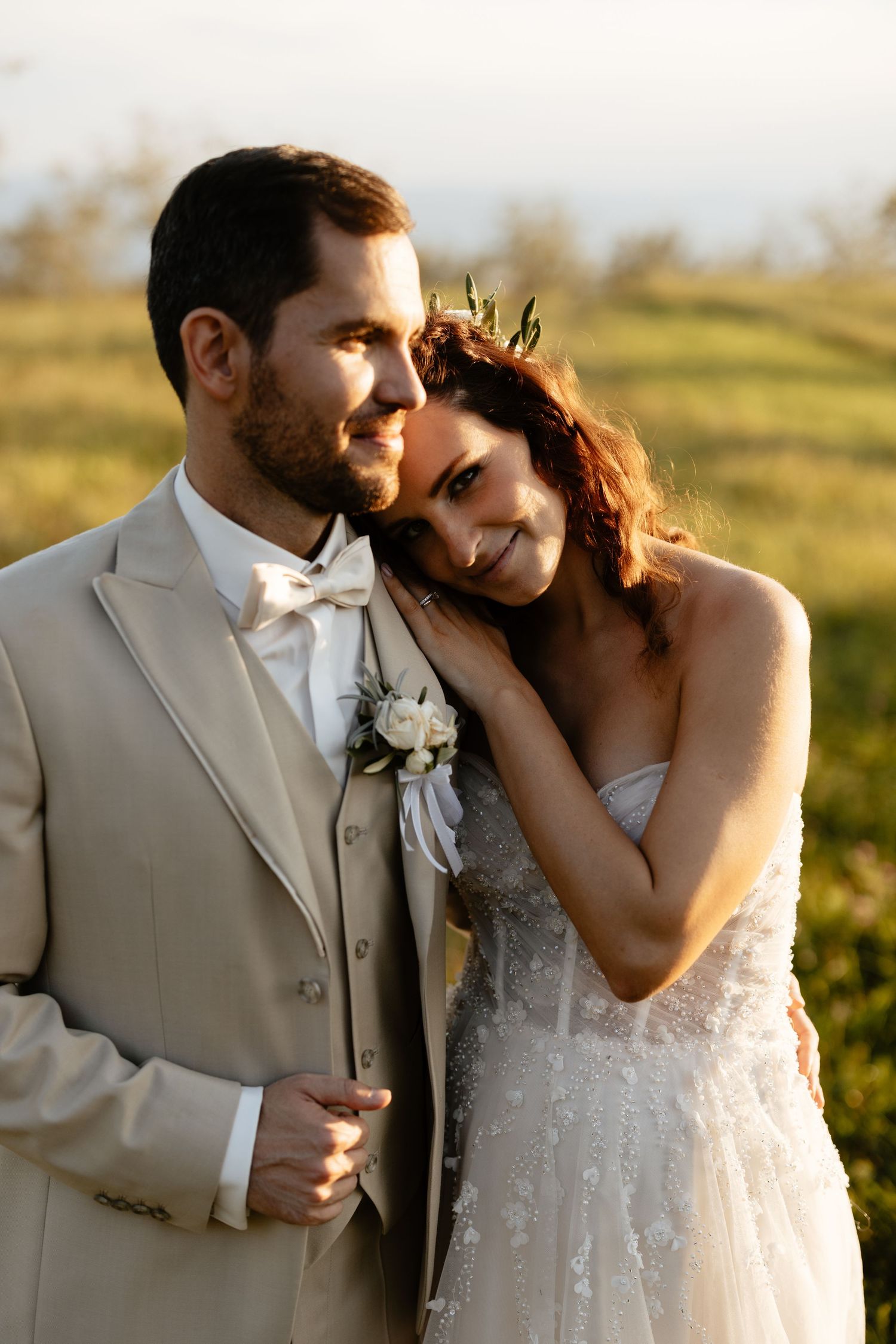 A romantic wedding couple shares intimate moments at sunset in a field while wearing a tan suit and white dress.