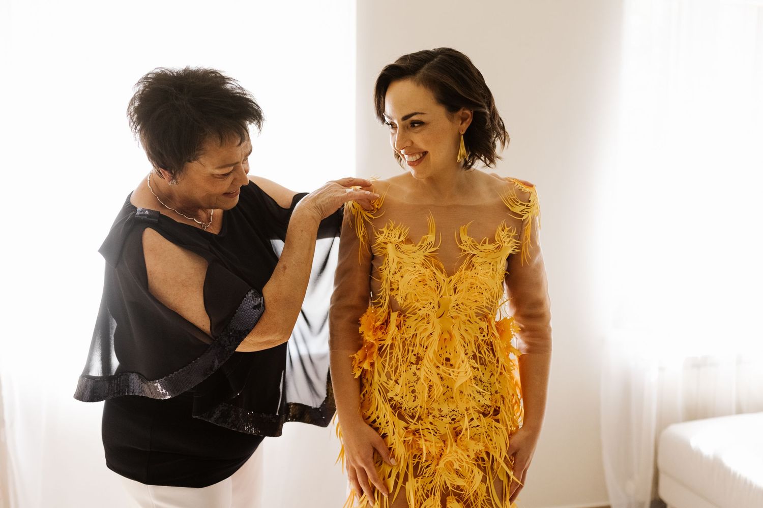 A stylist helps adjust a stunning yellow lace evening gown during a dress fitting session in a bright room.