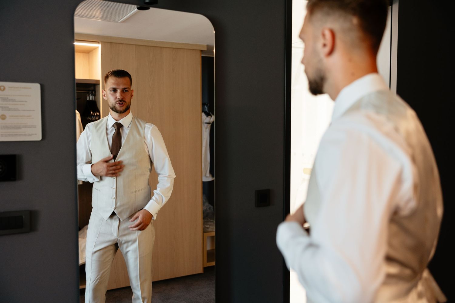 A groom adjusts his tie while getting ready in a bright hotel room with natural lighting.