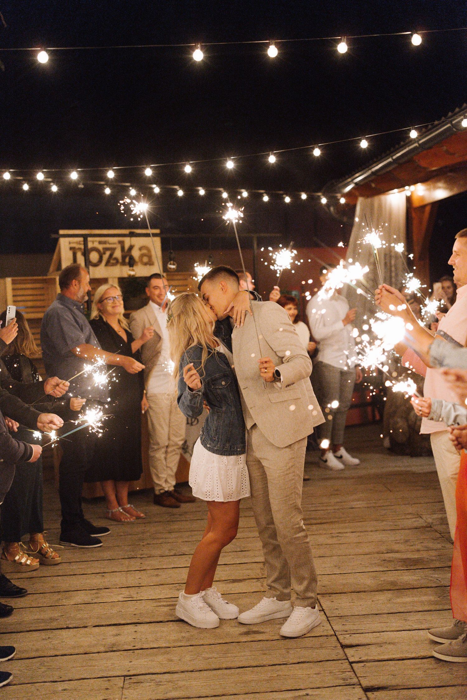 Wedding guests holding sparklers create a magical nighttime celebration on a wooden deck under string lights.