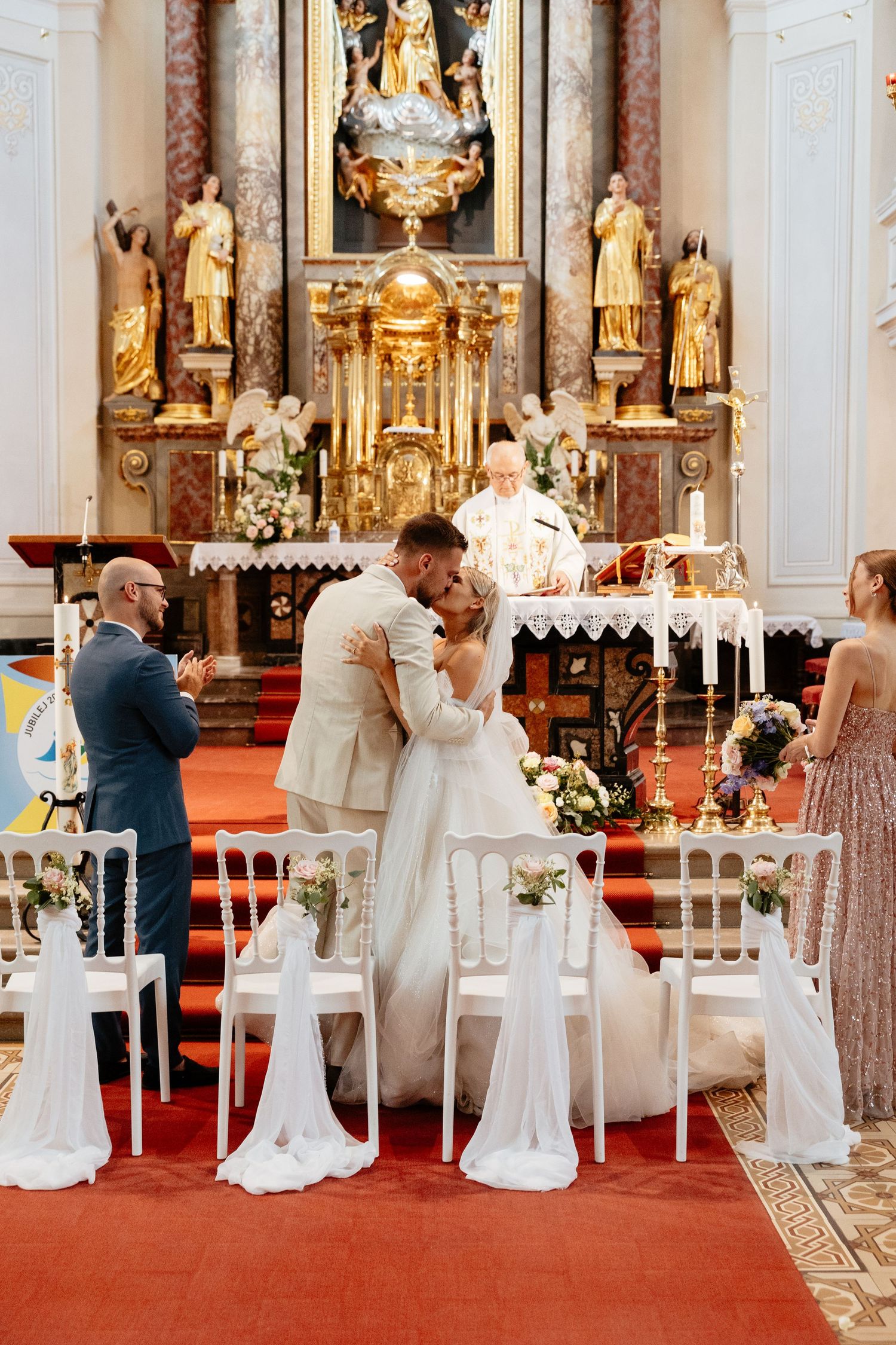 A wedding ceremony in an ornate church with gold altar decorations and white floral arrangements.