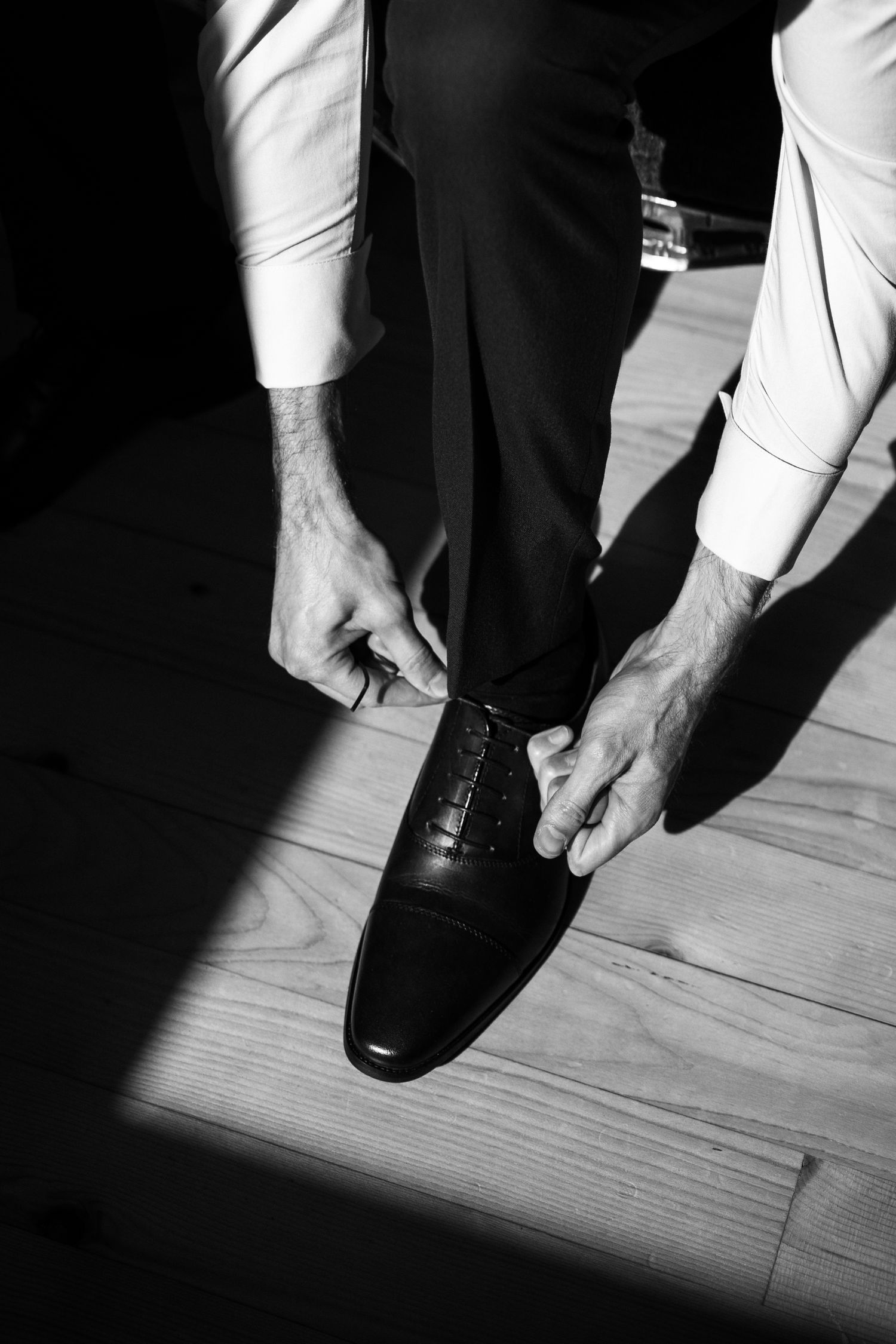 Black and white image of hands tying formal leather dress shoes on wooden floor in dramatic lighting.