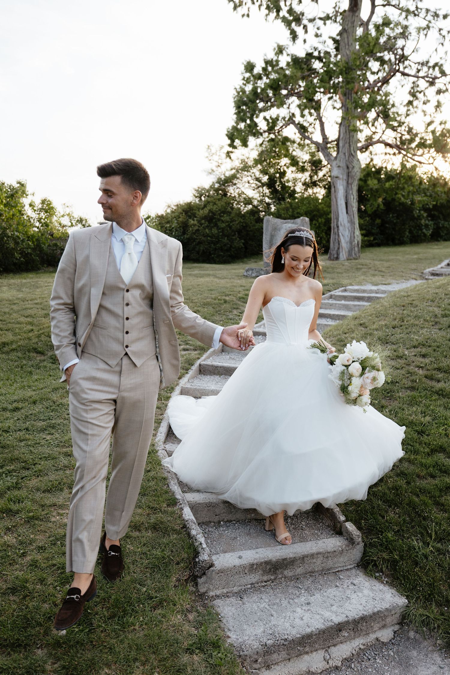 Couple descends outdoor stone steps playfully, bride's dress flowing in the breeze.
