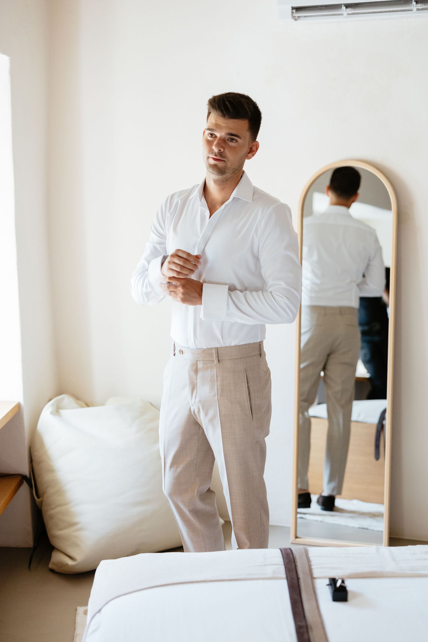 A groom in a crisp white shirt and light pants checks his appearance in a bedroom mirror.
