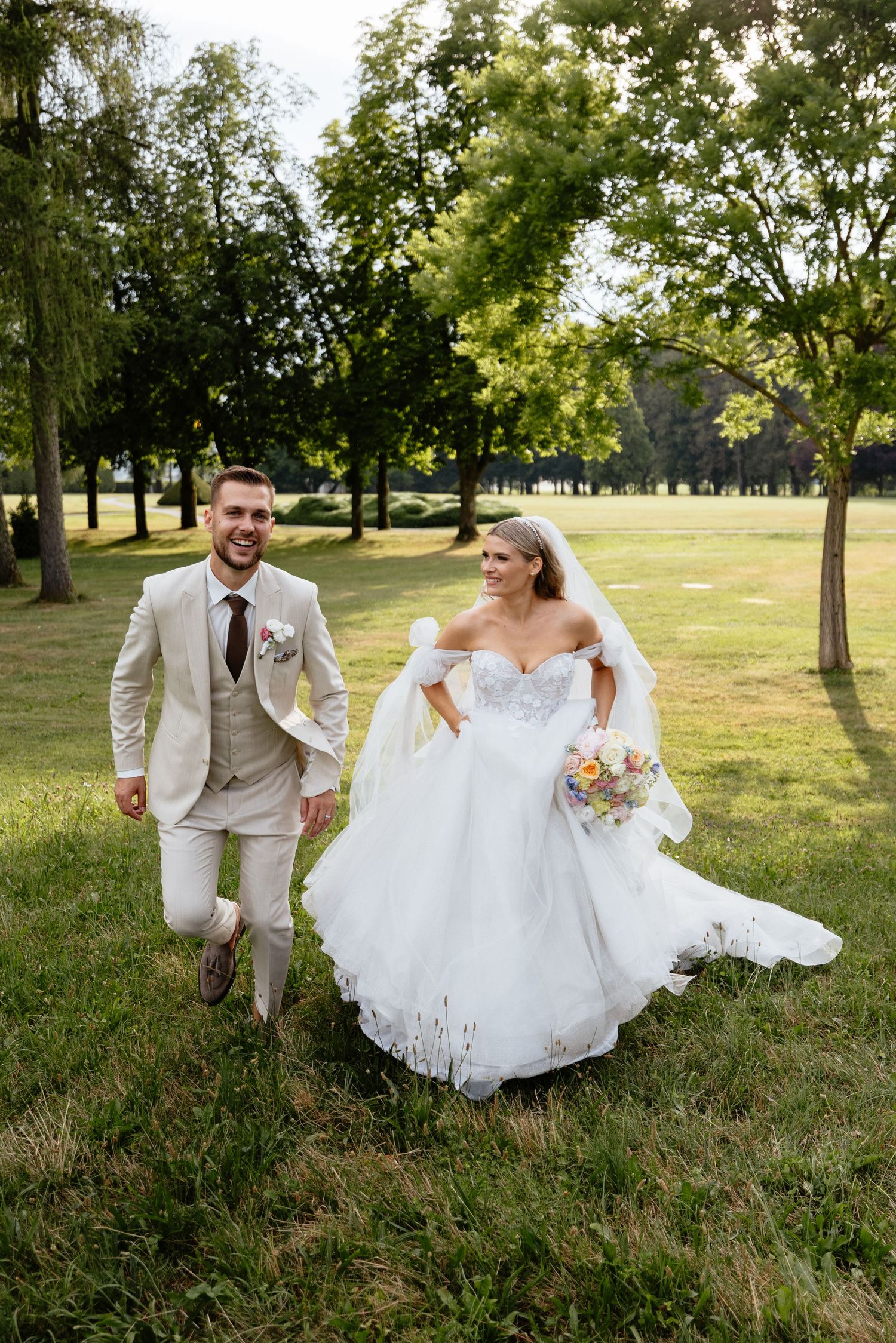 A couple in wedding attire runs playfully through a sunlit park with green grass and trees in the background.