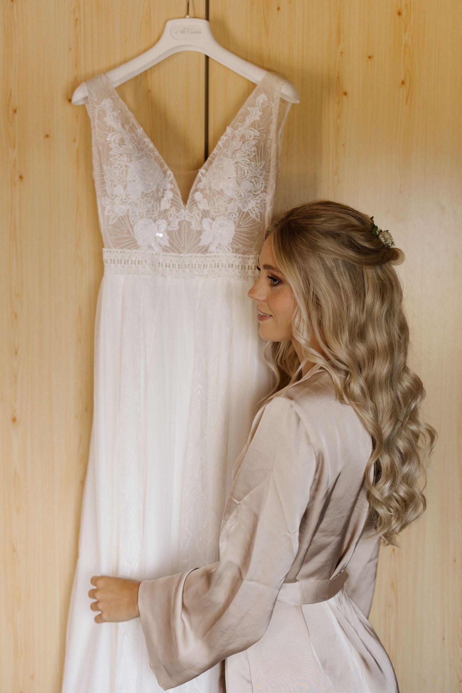 A white lace wedding dress hangs on a wooden wall while someone in a robe admires it.